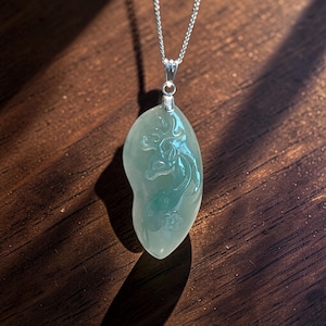 May include: A light green jade pendant with a carved design of a mythical creature, suspended from a silver chain. The pendant has a smooth, polished surface and is set against a dark wooden background.