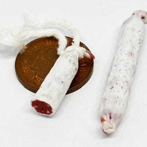 May include: Two small, white sausages with red ends and a coin. One sausage is tied with white string and rests on a brown coin. The other sausage is next to it. The sausages have a white, speckled exterior.