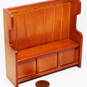 May include: Miniature wooden bench with a warm, reddish-brown finish. The bench features a back with vertical slats, a seat, and a lower section with three panels. A penny is next to the bench for scale, suggesting a dollhouse or model furniture item.