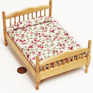 May include: A miniature wooden bed with a floral patterned comforter and pillow. The bed frame is light brown, and the bedding features a white background with pink and red roses. A penny is next to the bed for scale.