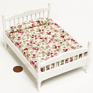 May include: A miniature white wooden bed with a floral patterned mattress and pillow. The bed frame has a headboard and footboard with vertical spindles. The bedding features a cream background with pink and red roses and green leaves.