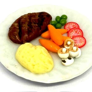 Peut inclure: Une assiette miniature de nourriture comprenant un steak grillé, de la purée de pommes de terre, des carottes, des petits pois, des tranches de tomates et des champignons. Le repas est présenté sur une assiette blanche.