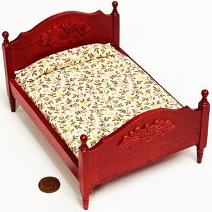 Puede incluir: Una cama de madera en miniatura pintada de rojo oscuro. El cabecero y el piecero presentan diseños florales tallados. La cama está hecha con una colcha estampada con flores. Una moneda está al lado de la cama para la escala.