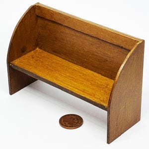 May include: A miniature wooden bench with a curved back and a flat seat. The bench is made of stained wood, with a warm, golden-brown hue. A penny is placed next to the bench for scale.