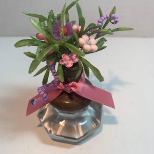 May include: A miniature floral arrangement in a clear glass container. The arrangement features green leaves, purple and pink flowers, and a pink ribbon. The container has a brown top and a faceted, hexagonal base.