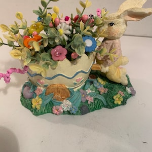 Puede incluir: Una figura decorativa de Pascua con un conejito color crema sosteniendo un huevo agrietado lleno de flores y vegetación coloridas. La base es un área cubierta de hierba con más flores y una pequeña puerta de madera.