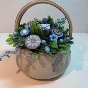May include: A ceramic basket with a handle, filled with artificial greenery, blue button flowers, and beaded accents. The basket has a light brown glaze with blue and green leaf patterns.