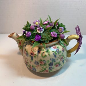 Puede incluir: Una tetera decorativa con un estampado floral en azul, rosa y rojo. La tetera está llena de flores artificiales en tonos morados y verdes. Una cinta morada con lunares blancos está atada al asa.