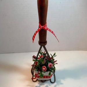 May include: A decorative floral arrangement featuring a vintage-style wooden handle with a red ribbon. The arrangement includes a small white planter with red and white gingham accents, filled with greenery and red floral elements.