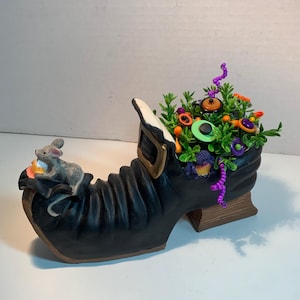 May include: A black shoe-shaped planter with a gold buckle, featuring a small gray mouse figurine perched on the toe. The interior is filled with green foliage, purple beaded accents, and various colorful buttons in orange, green, and purple.