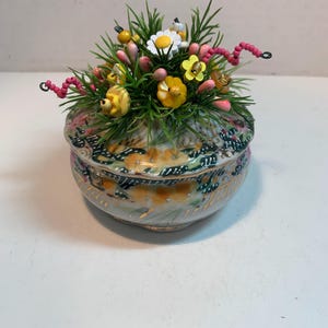 May include: A decorative ceramic trinket box with a lid, adorned with a floral arrangement. The box features a painted floral design in shades of yellow, green, and gold. The arrangement on top includes yellow, white, and pink flowers with green foliage.