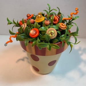 May include: A decorative flower pot with a beige base and brown polka dots. The pot is filled with faux flowers made from buttons in shades of orange, yellow, and tan, with green leaves and orange beaded accents. The pot has a striped rim.