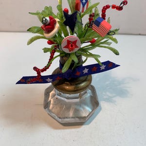 May include: A decorative arrangement featuring a clear glass knob base with a brass-colored top. The arrangement includes green foliage, red beaded accents, and patriotic-themed embellishments such as a small American flag, a Puerto Rican flag, and star-shaped buttons.