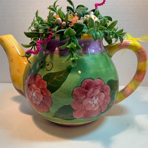 May include: A ceramic teapot featuring a green body with painted pink flowers and dark green leaves. The yellow spout has orange cross-stitch patterns, and the handle has yellow and pink stripes. It is filled with green faux foliage and pink beads.