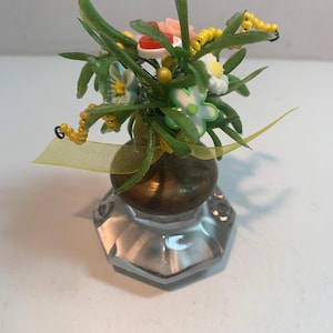 May include: A miniature floral arrangement in a vintage-style glass vase. The arrangement features green leaves, yellow beaded accents, and small, colorful flowers. A yellow ribbon is tied around the vase. The vase has a clear, faceted base.