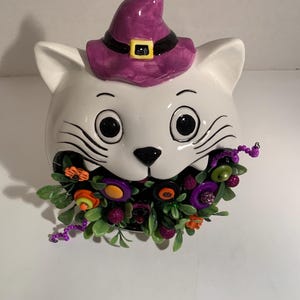 May include: A white ceramic cat head figurine wearing a purple witch hat with a black band and yellow buckle. The cat has black whiskers and eyes, and its open mouth is filled with a decorative arrangement of green leaves, purple beads, and buttons.
