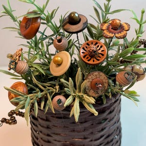 May include: A decorative arrangement of button flowers in a dark brown woven basket. The button flowers are various shapes and colors, including brown, tan, and orange. Green faux foliage adds a natural touch to the arrangement.