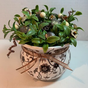 May include: A decorative floral arrangement in a white ceramic pot with a brown floral pattern and a brown ribbon. The arrangement features green leaves, decorative beads, and various embellishments.