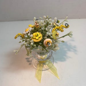 May include: A small, clear glass vase holds a miniature floral arrangement. The arrangement features yellow button-like flowers, white berries, and green stems. A yellow ribbon is tied around the base of the vase, adding a decorative touch. The overall aesthetic is delicate and charming.