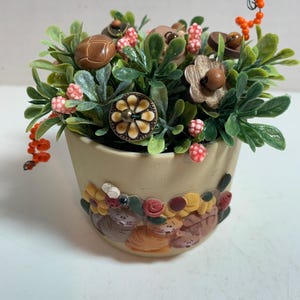 May include: A decorative ceramic pot filled with faux greenery, buttons, and berries. The pot is beige with a floral and cat relief design around the base. Buttons are brown, beige, and cream-colored, with some resembling flowers.