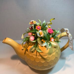 May include: A yellow ceramic teapot with a textured honeycomb pattern on the exterior, filled with artificial green foliage, small colorful flower embellishments, and a small bee figure. A beige ribbon with a white daisy print is tied to the handle.
