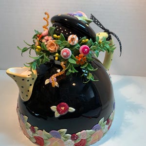 May include: A black ceramic teapot featuring a yellow spout with black polka dots and a yellow handle. The lid is adorned with green foliage, colorful buttons, and orange beads. A floral border in shades of pink, red, and green encircles the base.
