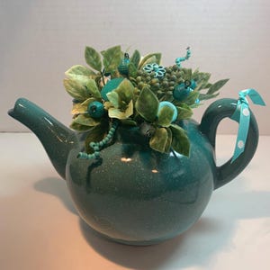 May include: Teal ceramic teapot with a speckled finish, filled with faux green leaves, turquoise beads, and decorative accents. A turquoise ribbon with white polka dots adorns the handle. A decorative floral arrangement.
