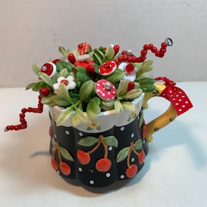 May include: A decorative ceramic mug with a black base adorned with white polka dots and red cherries. The mug is filled with artificial flowers, red berries, and button-like accents. The handle is yellow with a red and white polka dot ribbon.