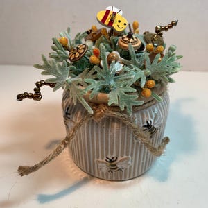 Puede incluir: Una maceta decorativa de cerámica con temática de abejas. La maceta beige presenta rayas verticales y diseños de abejas en relieve. Está adornada con un lazo de cuerda y llena de vegetación artificial, botones de madera y un acento de abeja de madera. La maceta mide unos 10 cm de alto.
