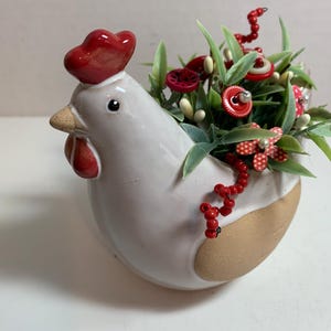 May include: A white ceramic hen-shaped planter with a red comb and wattle. The planter is filled with green foliage, red buttons, and beaded accents. The base of the planter is tan.