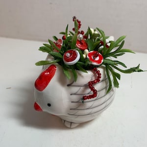 May include: A ceramic hen-shaped planter, white with gray lines and red accents, filled with faux greenery, red berries, and decorative red and white buttons. The planter has a small black eye and a red comb.