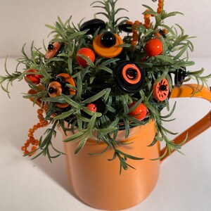 May include: An orange metal pitcher filled with green artificial foliage and decorative buttons in shades of black and orange. The buttons are layered and attached to the plant stems, interspersed with small orange beads on thin wire.