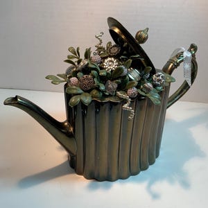 May include: A dark bronze-colored teapot with a fluted body and a partially open lid. The interior is filled with green artificial foliage and various metallic and pearl-toned decorative buttons and beads. A silver ribbon is tied to the handle.
