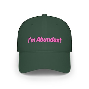 May include: A dark forest green baseball cap featuring the text "I'm Abundant" embroidered in a bright pink font across the front center. The cap has a structured crown, a curved brim, and a button at the top center of the fabric panels.