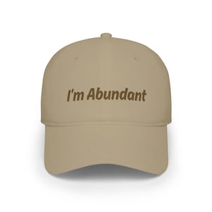 May include: A beige baseball cap featuring the text I'm Abundant embroidered in dark brown thread across the front panel. The cap has a structured crown, a curved brim, and a button at the top center, providing a simple and clean design.