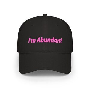 May include: A black baseball cap featuring the text I'm Abundant embroidered in a bright pink font across the front panel. The cap has a structured crown, a curved brim, and a button at the top center, presenting a clean and simple design.