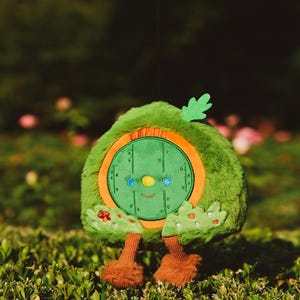 Puede incluir: Un juguete de peluche verde con forma de agujero de hobbit con una puerta redonda y una cara sonriente. El juguete tiene pies peludos marrones y está sentado en un lecho de hierba verde.