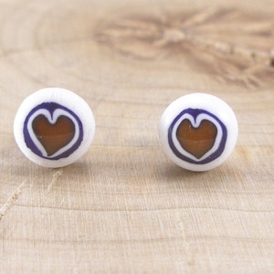 May include: A pair of white ceramic stud earrings with a brown heart design outlined in purple.