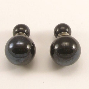 Puede incluir: Un par de pendientes de botón esféricos de color gris oscuro. Cada pendiente presenta una bola grande y brillante conectada a una bola más pequeña con un poste de metal plateado. Diseño sencillo y elegante.