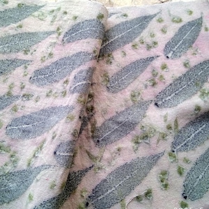 May include: Textile fabric featuring a repeating pattern of large, grey leaf imprints on a pink and white background. Small green floral accents are scattered throughout the design. The fabric is folded, showing the pattern's continuity.