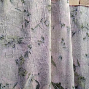 May include: Close-up of a light-coloured fabric with a botanical print. The fabric has a textured appearance with a pattern of green leaves and small floral elements. The folds of the fabric are visible, creating depth and shadow.