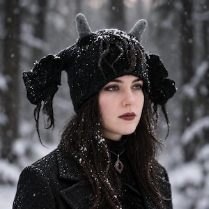Peut inclure: Un bonnet noir texturé doté de deux cornes grises feutrées dressées et de deux larges appendices latéraux noirs tombants aux bords effilochés. Le bonnet est recouvert d'une légère couche de neige blanche, créant un contraste avec le tissu sombre.