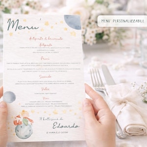 Puede incluir: Una tarjeta de menú con texto en italiano, con una ilustración de acuarela de un planeta y un personaje. La palabra "Menu" está en la parte superior. La tarjeta está sostenida por manos, con un fondo borroso de una mesa.