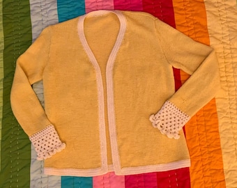 May include: A yellow knitted cardigan with white crocheted trim on the cuffs and down the front. The cardigan is laid flat on a colorful striped fabric.