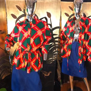 May include: A vintage-style outfit featuring a green, red, and yellow plaid shirt with a matching belt, paired with a blue skirt. The shirt has long sleeves and a collared neckline. The outfit is displayed on a mannequin.