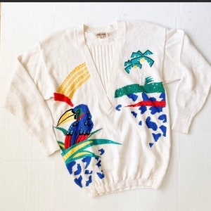 Op de afbeelding: Witte gebreide trui met een V-hals en een kleurrijk geborduurd tropisch ontwerp. Het ontwerp bevat een toekan, palmbomen en abstracte vormen in rood, geel, blauw en groen.