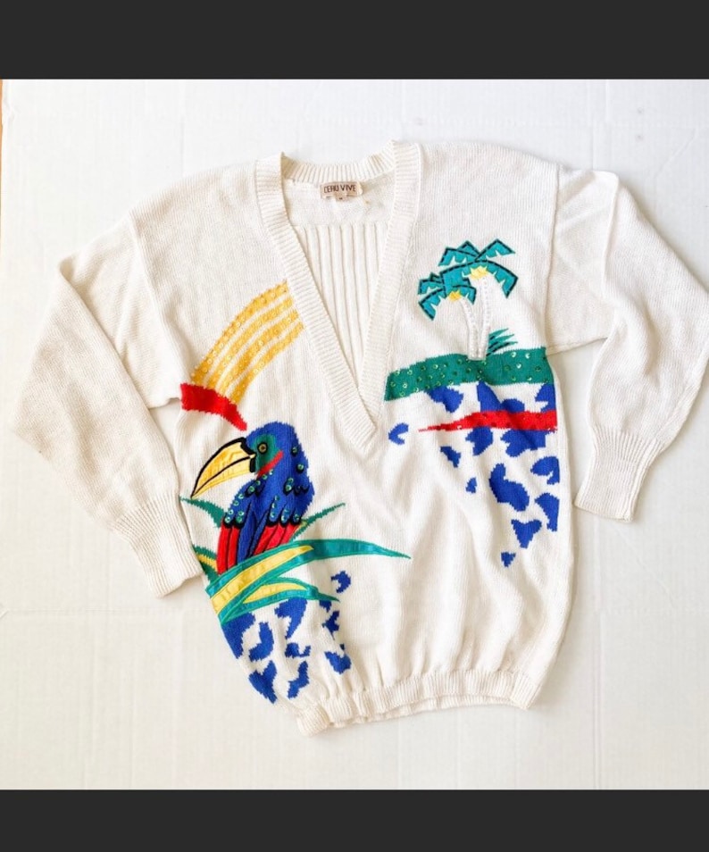 Op de afbeelding: Witte gebreide trui met een V-hals en een kleurrijk geborduurd tropisch ontwerp. Het ontwerp bevat een toekan, palmbomen en abstracte vormen in rood, geel, blauw en groen.