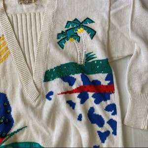 Op de afbeelding: Een witte gebreide trui met een V-hals en kleurrijke geborduurde ontwerpen. De trui heeft palmbomen, een rood-wit-blauw geometrisch patroon en een blauw-witte dierenprint. De trui is gemaakt van een zacht, comfortabel materiaal.