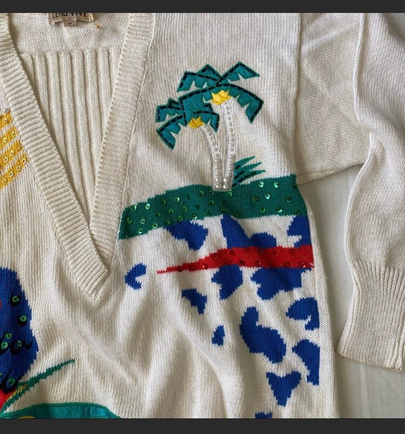 Op de afbeelding: Een witte gebreide trui met een V-hals en kleurrijke geborduurde ontwerpen. De trui heeft palmbomen, een rood-wit-blauw geometrisch patroon en een blauw-witte dierenprint. De trui is gemaakt van een zacht, comfortabel materiaal.
