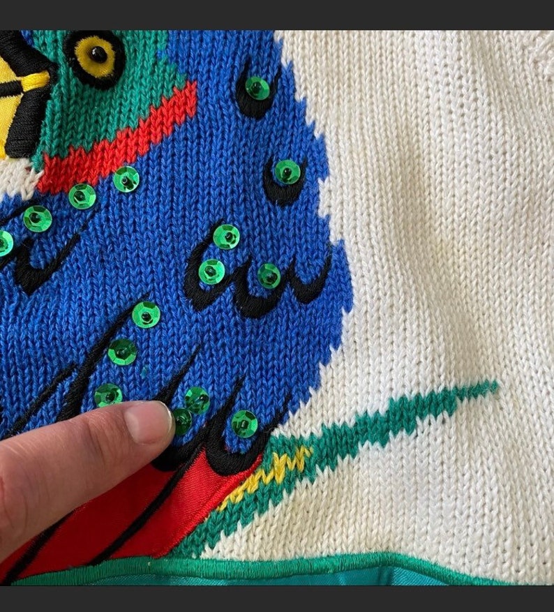 Op de afbeelding: Een witte gebreide trui met een kleurrijk papegaai-ontwerp. De papegaai heeft groene, blauwe, rode en gele veren. De ogen van de papegaai zijn zwart met groene pailletten. De trui heeft een groene en gele rand.